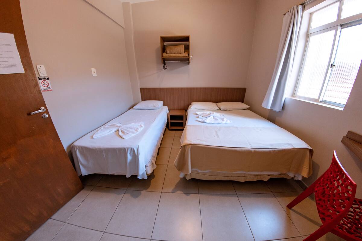 Una habitación pequeña y limpia con dos camas, un estante de madera encima, una silla roja y una ventana con cortinas.