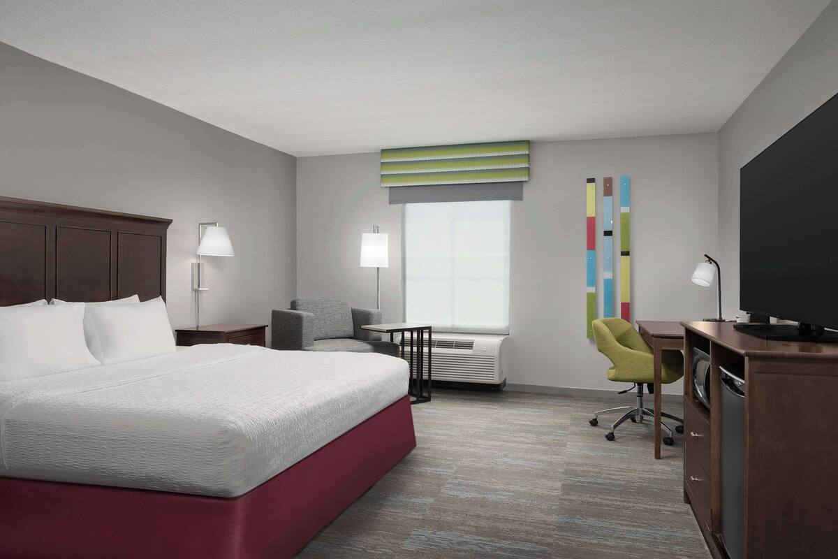 A hotel room with a large bed, a desk, a chair, a television, and a window with blinds.