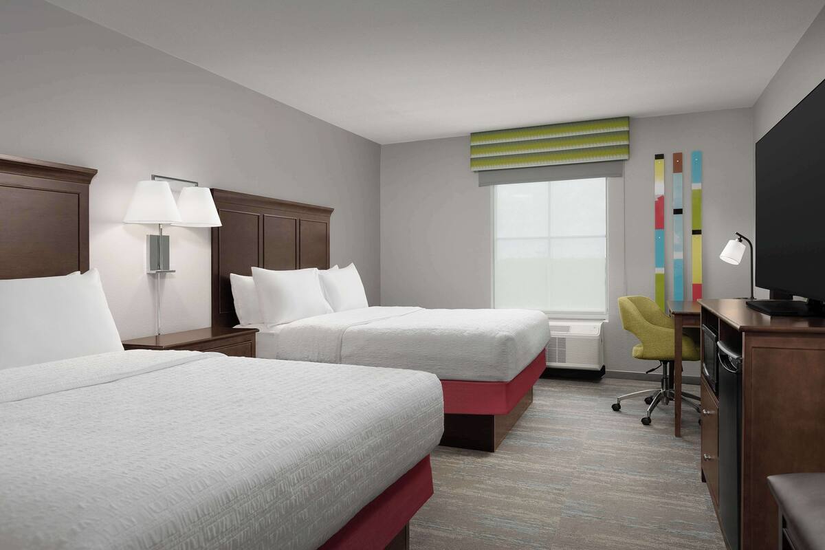 A hotel room with two beds, a desk, a chair, a TV, and a window with blinds.