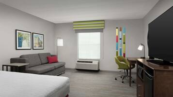 Habitación de hotel con cama, sofá gris, escritorio con silla, televisor de pantalla plana grande, ventana con persianas y cuadros en la pared.