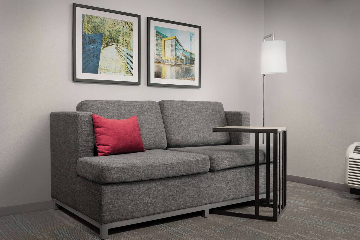 A modern living room with a grey sofa, a red pillow, a small side table, and two framed artworks on the wall.