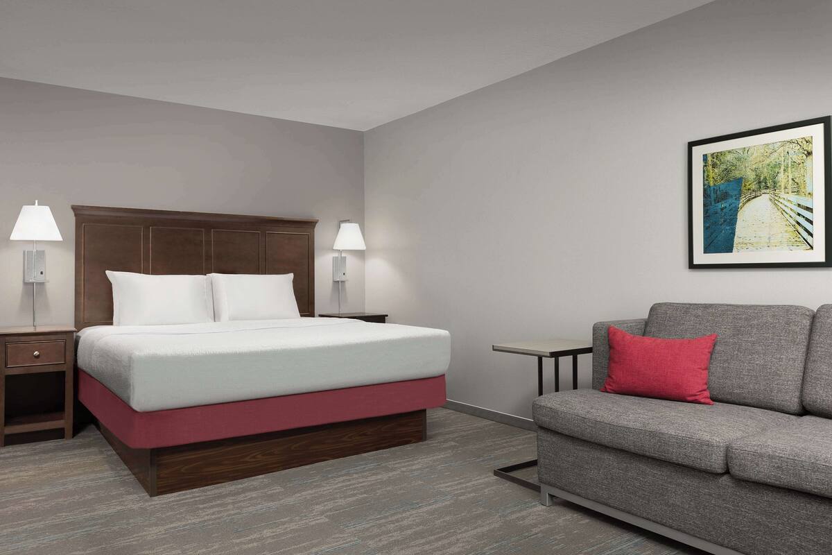 A hotel room with a bed, a grey sofa, a nightstand, and a wall-mounted lamp.