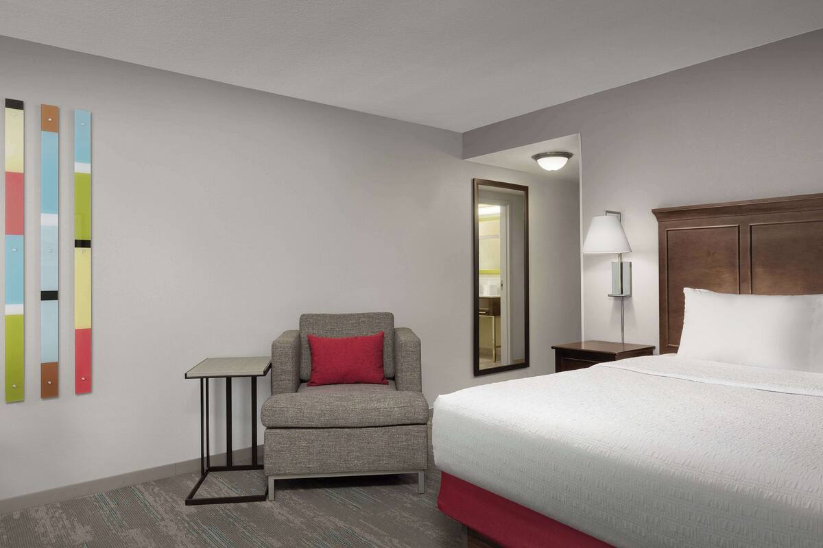 A hotel room with a bed, a chair, a small table, a lamp, and a mirror.
