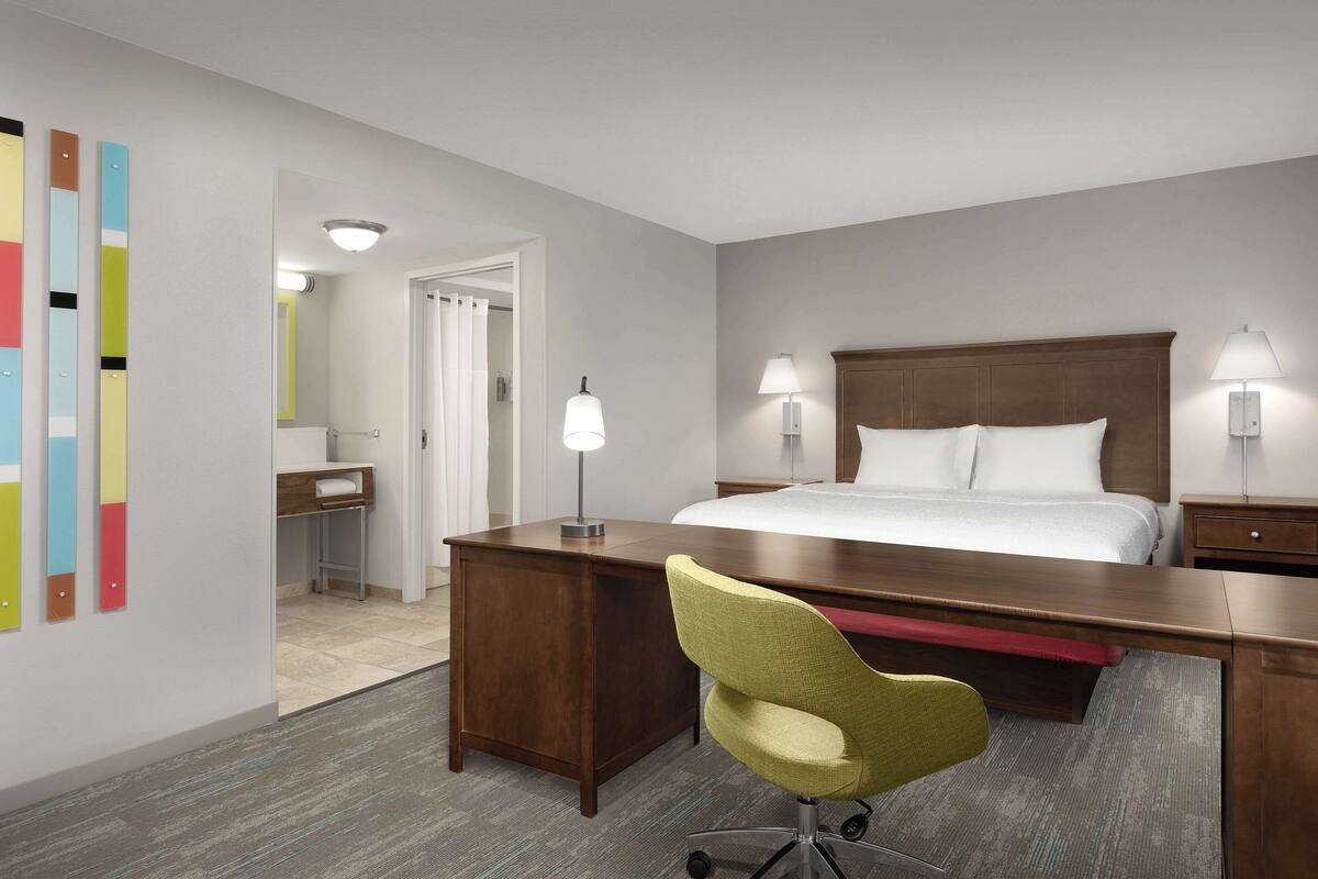 A hotel room with a large bed, two bedside lamps, a desk with a chair, a mirror, and a door leading to a bathroom.