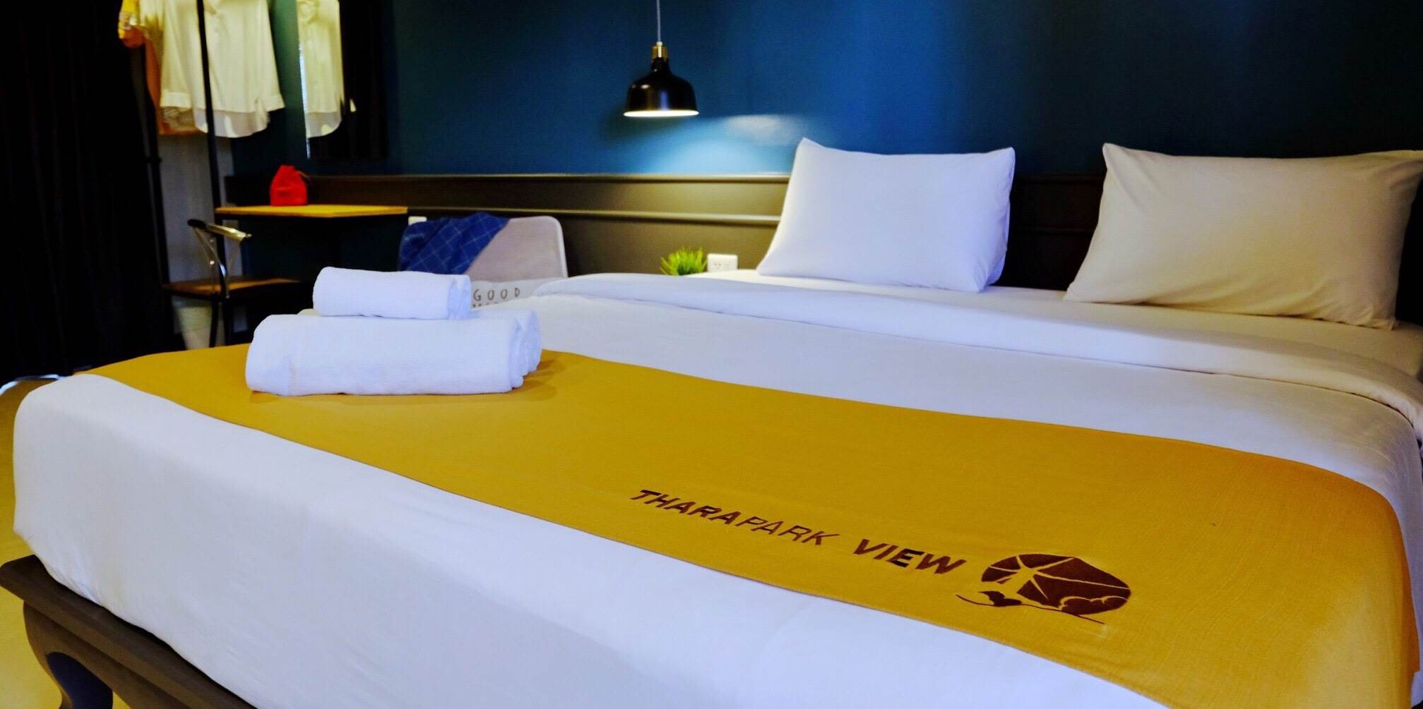 A hotel room with a bed featuring a THARAPARK VIEW logo, white bedding, and a yellow accent.