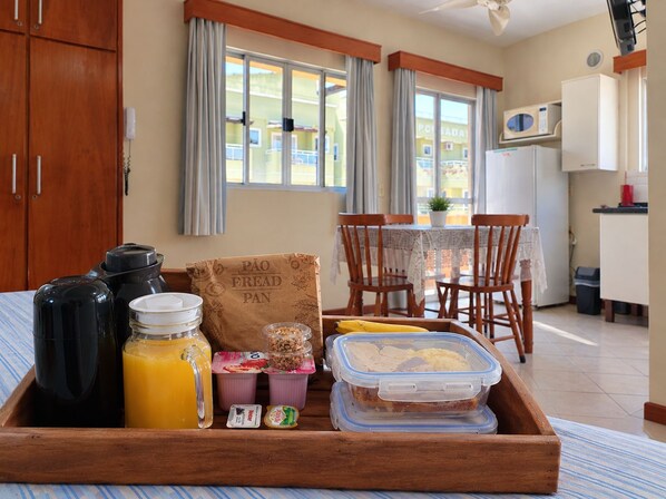 Bandeja de desayuno con una botella, jugo, yogur y una banana en una habitación de hotel con una mesa de comedor, sillas y vistas a edificios.