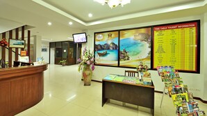 A hotel reception area with a wooden reception desk, a television screen, a large wall clock, and a time table for tourist information. - Lada Krabi Residence (Krabi)