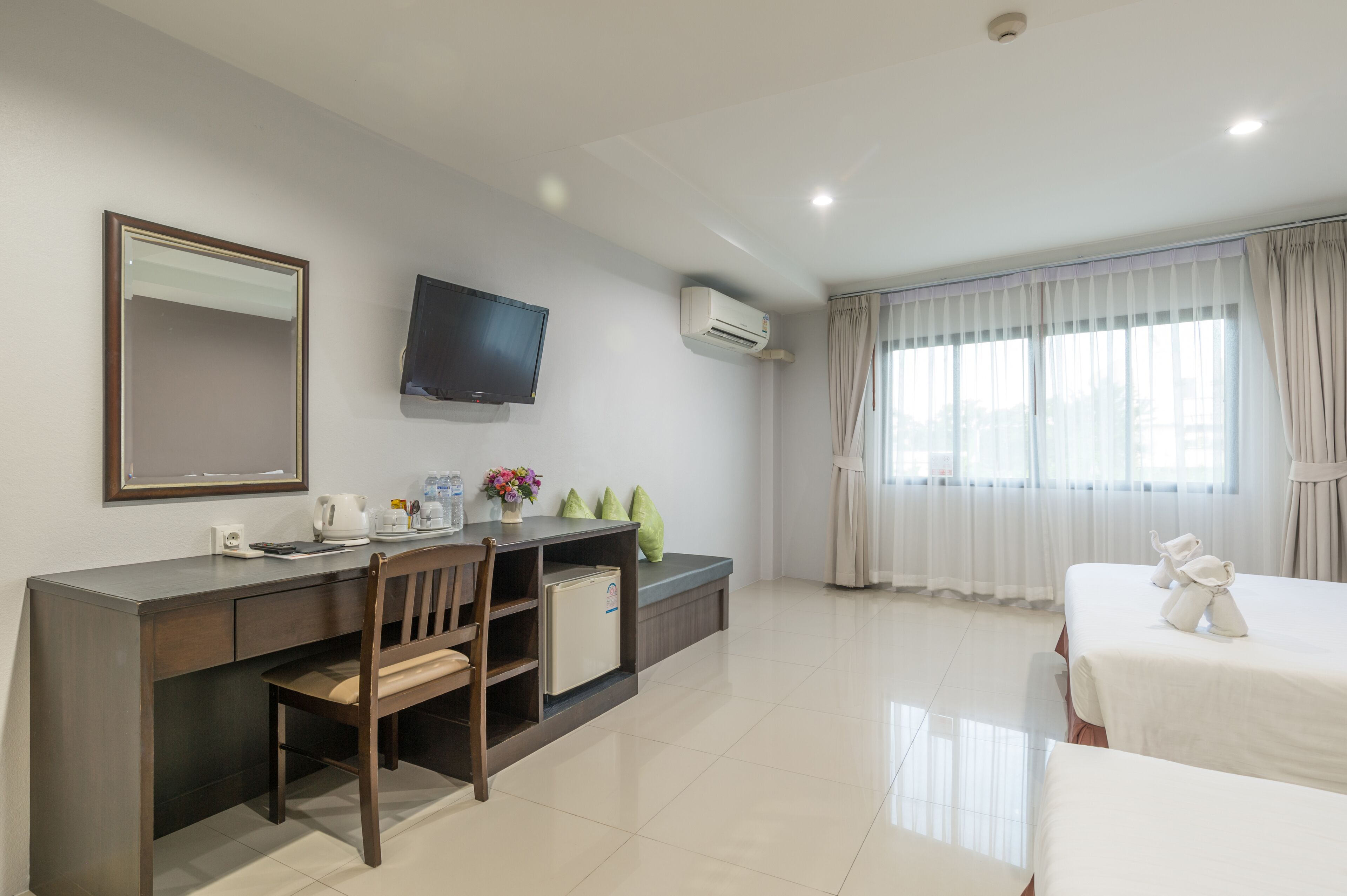 A hotel room with a bed, a desk with a chair, a TV, an air conditioner, and a window with curtains.