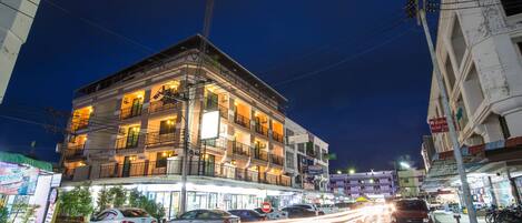Un edificio de varios pisos con balcones, una calle con coches y letreros iluminados.