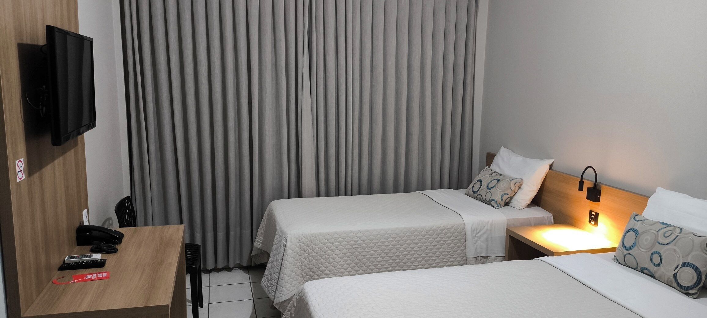 Quarto de hotel com duas camas, uma TV fixada na parede, uma escrivaninha com telefone e controle remoto, e uma cortina.