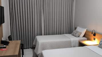 Quarto de hotel com duas camas, uma TV fixada na parede, uma escrivaninha com telefone e controle remoto, e uma cortina.