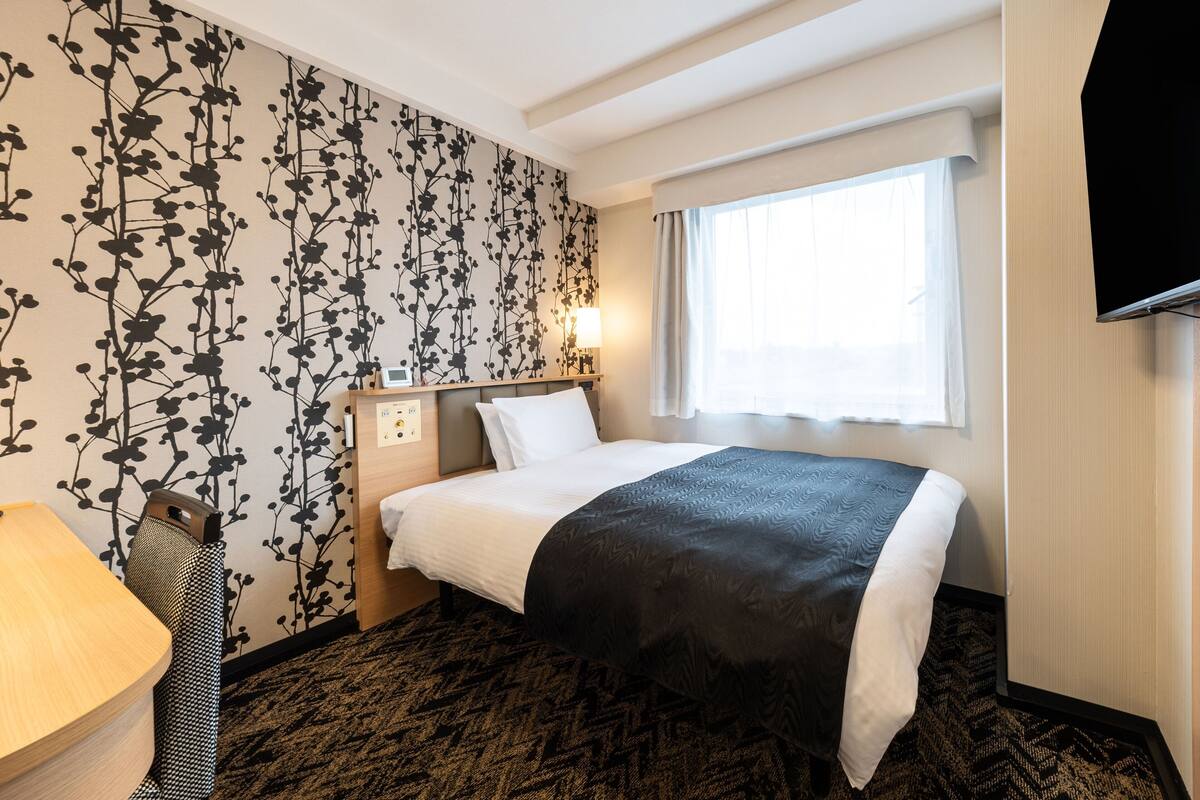 A hotel room with a bed, a bedside table, a lamp, a TV, and a window with curtains.