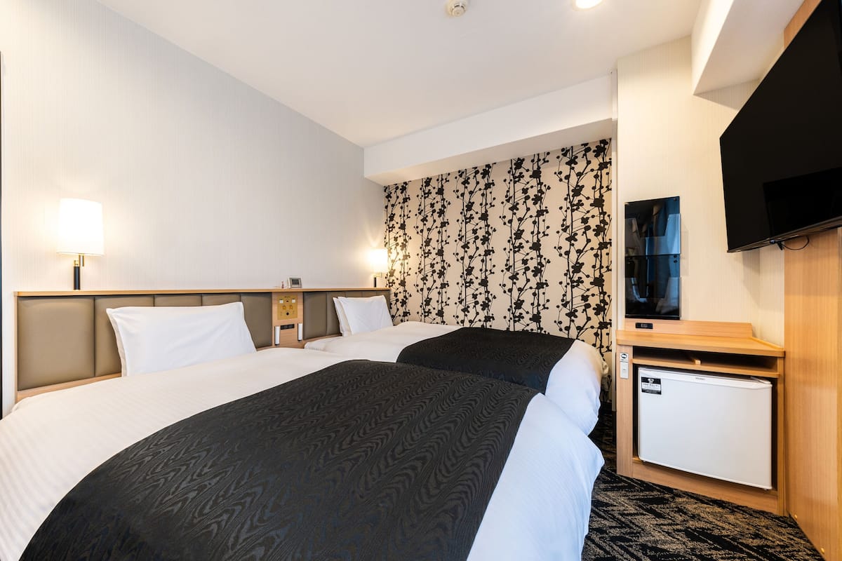 A hotel room with two beds, a small desk, a television, and a wall with a floral pattern.