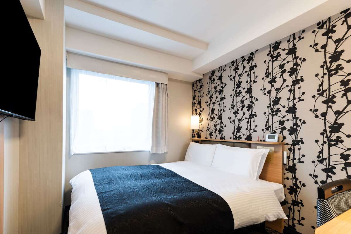 A hotel room with a bed, a TV, a window with curtains, a wall with a floral pattern, and a bedside table with a lamp.