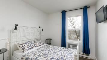 A bedroom with a white metal bed frame, a patterned bedspread, a bedside table with a lamp, a window with blue curtains, and a wall-mounted TV.