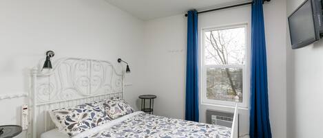 A bedroom with a white metal bed frame, a patterned bedspread, a bedside table with a lamp, a window with blue curtains, and a wall-mounted TV.