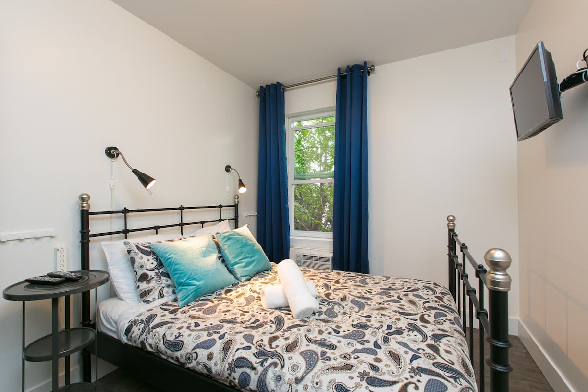 Una cama bien hecha con un edredón estampado, una mesita de noche con una lámpara y una ventana con cortinas.