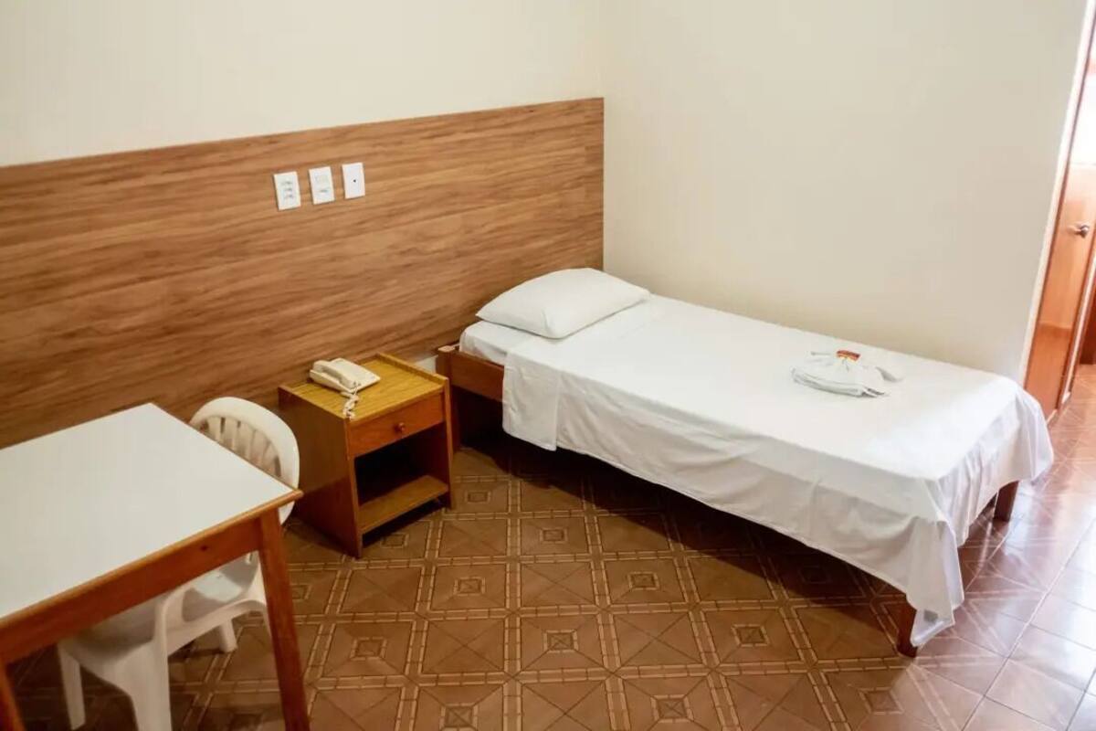 Una habitación pequeña y limpia con tablero de cama de madera, una cama individual con ropa de cama blanca, una mesita de noche de madera con un teléfono, un escritorio blanco y una silla.