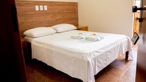 A neatly made bed with white linens and a wooden headboard, a small bedside table, and a tiled floor. - Hotel Almanara (Cuiabá)