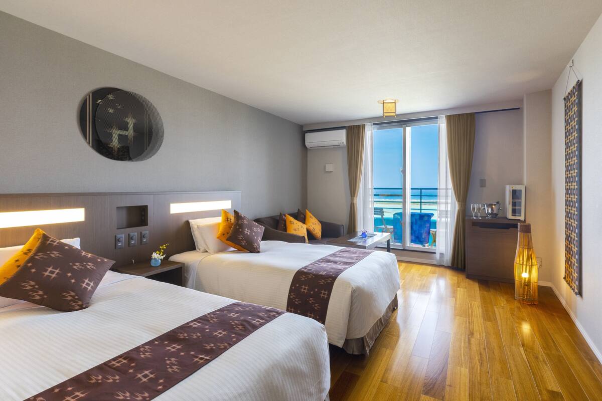Ett hotellrum med två sängar, ett stort fönster med gardiner, ett litet bord med en lampa och utsikt över havet.