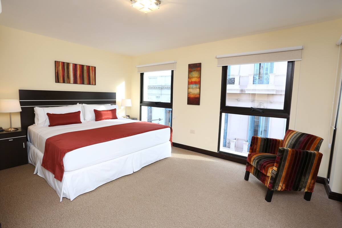 Ein Hotelzimmer mit einem großen Bett, zwei Nachttischen, einem Sessel und großen Fenstern mit Jalousien.