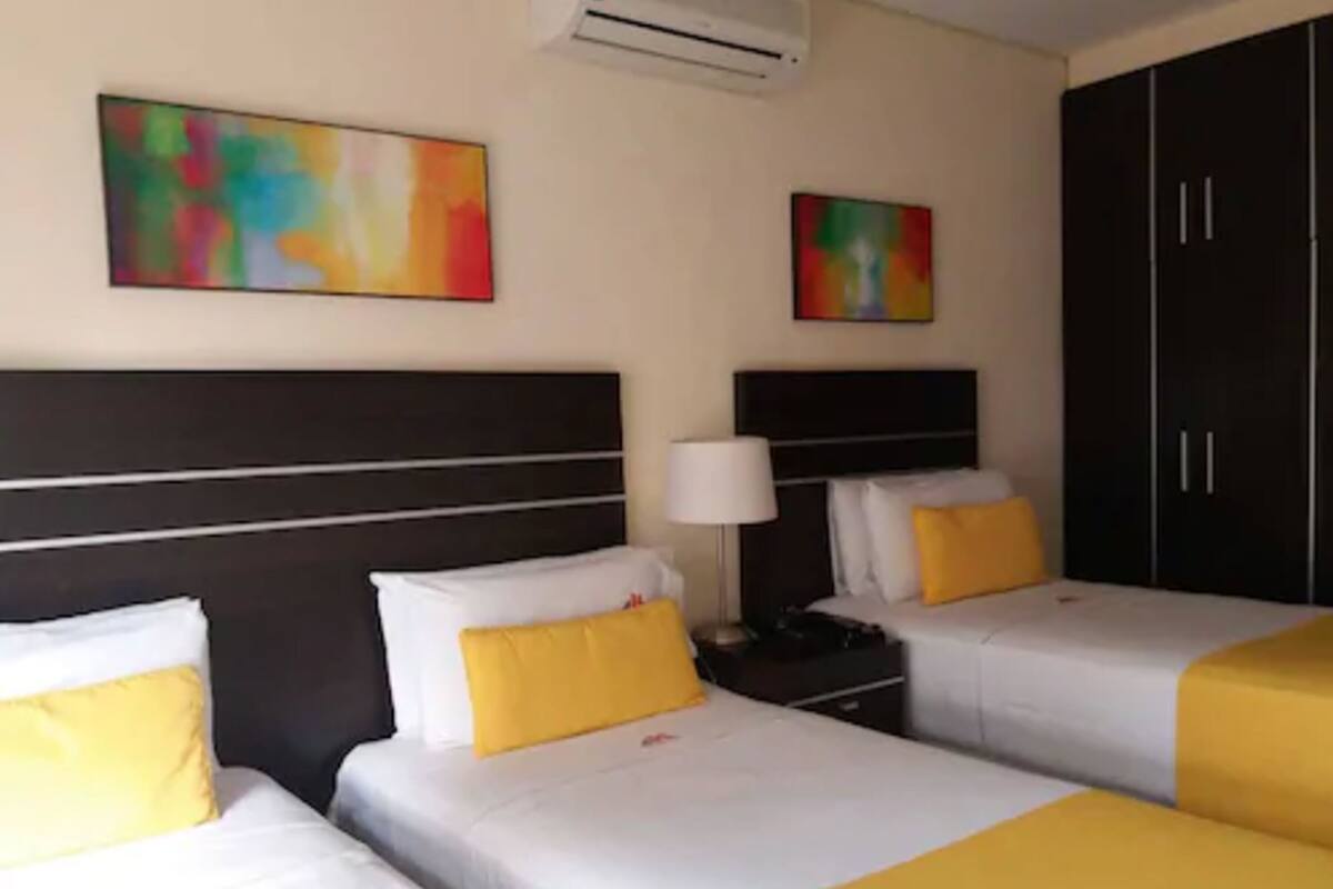 Ein Hotelzimmer mit zwei Betten, jeweils mit weißer Bettwäsche und gelben Akzenten. Zwei Wand-Klimaanlagen und zwei abstrakte Gemälde sind vorhanden.