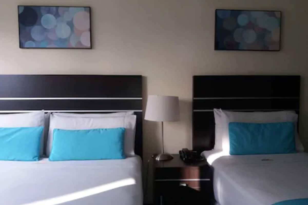 Ein Hotelzimmer mit zwei Betten, jeweils mit blauer und weißer Bettwäsche, einem Nachttisch mit Lampe sowie abstrakten Kunstwerken an den Wänden.