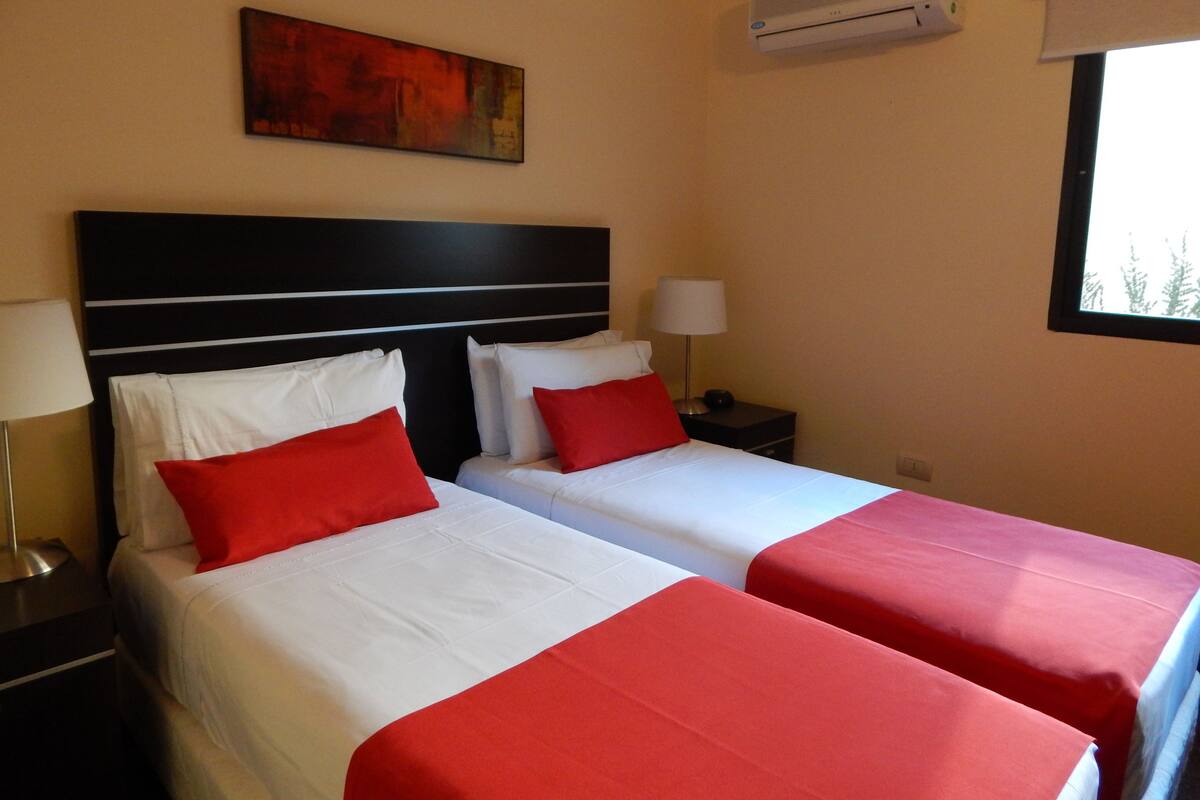 Ein Hotelzimmer mit zwei Betten, jeweils mit weißer Bettwäsche und roten Akzenten, einem schwarzen Kopfteil, einem Gemälde an der Wand, einer Klimaanlage und einem Fenster mit Blick.