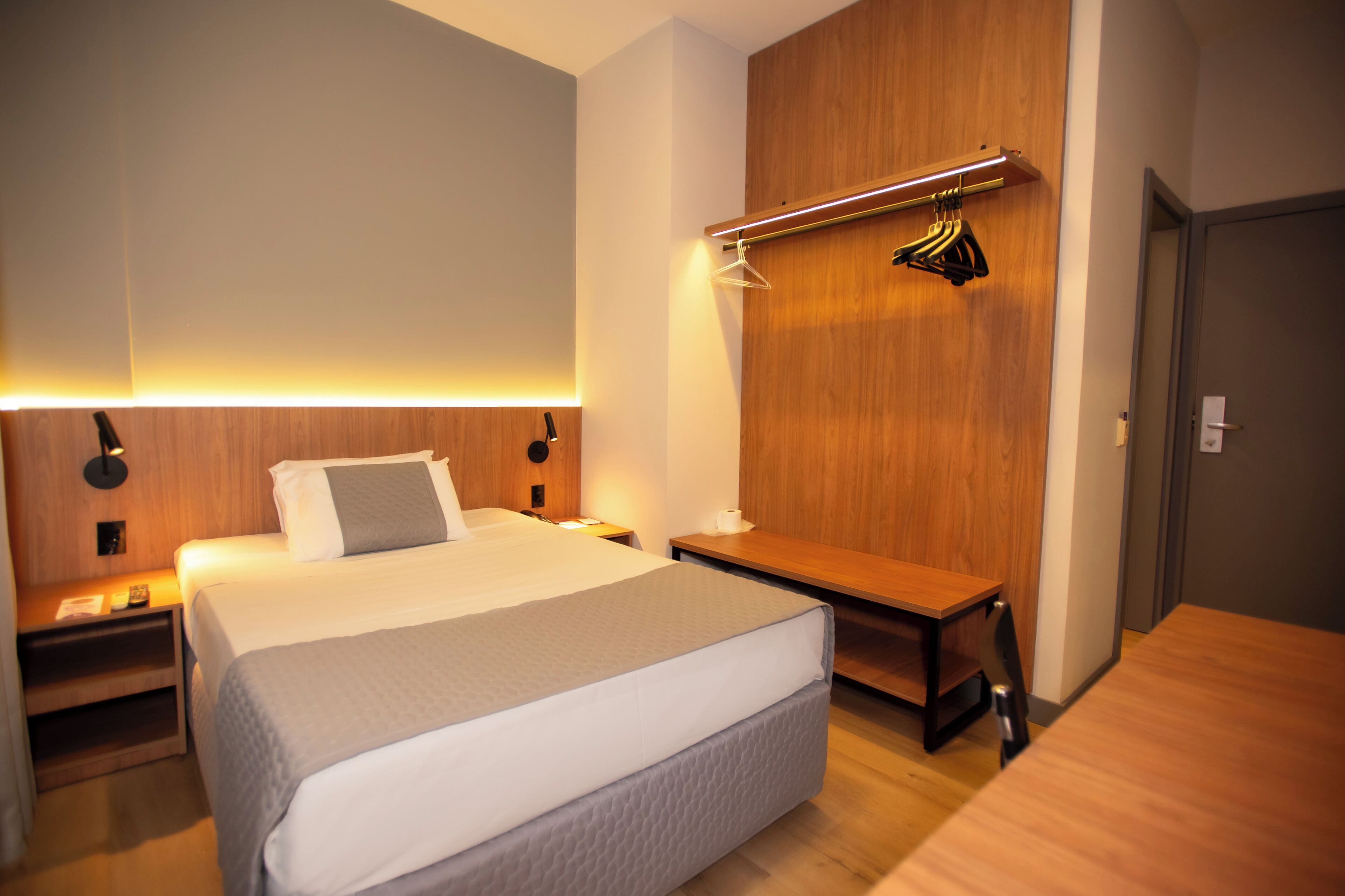 A modern hotel room with a large bed, bedside tables, a wooden wardrobe, and a desk with a chair.