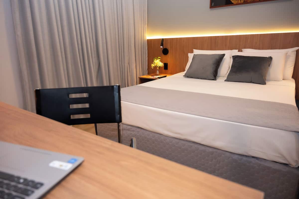 Una habitación de hotel con cama, escritorio con laptop, silla, mesita de noche con lámpara y ventana con cortinas.