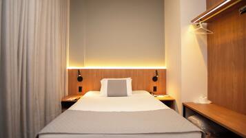 Quarto de hotel com cama bem arrumada, cabeceira de madeira, mesas de cabeceira com abajures, criado-mudo de madeira e um banco de madeira com luminária pendente.