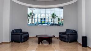 A lobby with two black leather sofas, a round table, and a large window with a view of a building and palm trees. - Delcas Hotel (Cuiabá)