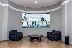 Um hall com dois sofás de couro preto, uma mesa redonda e uma janela grande com vista para um edifício e palmeiras.