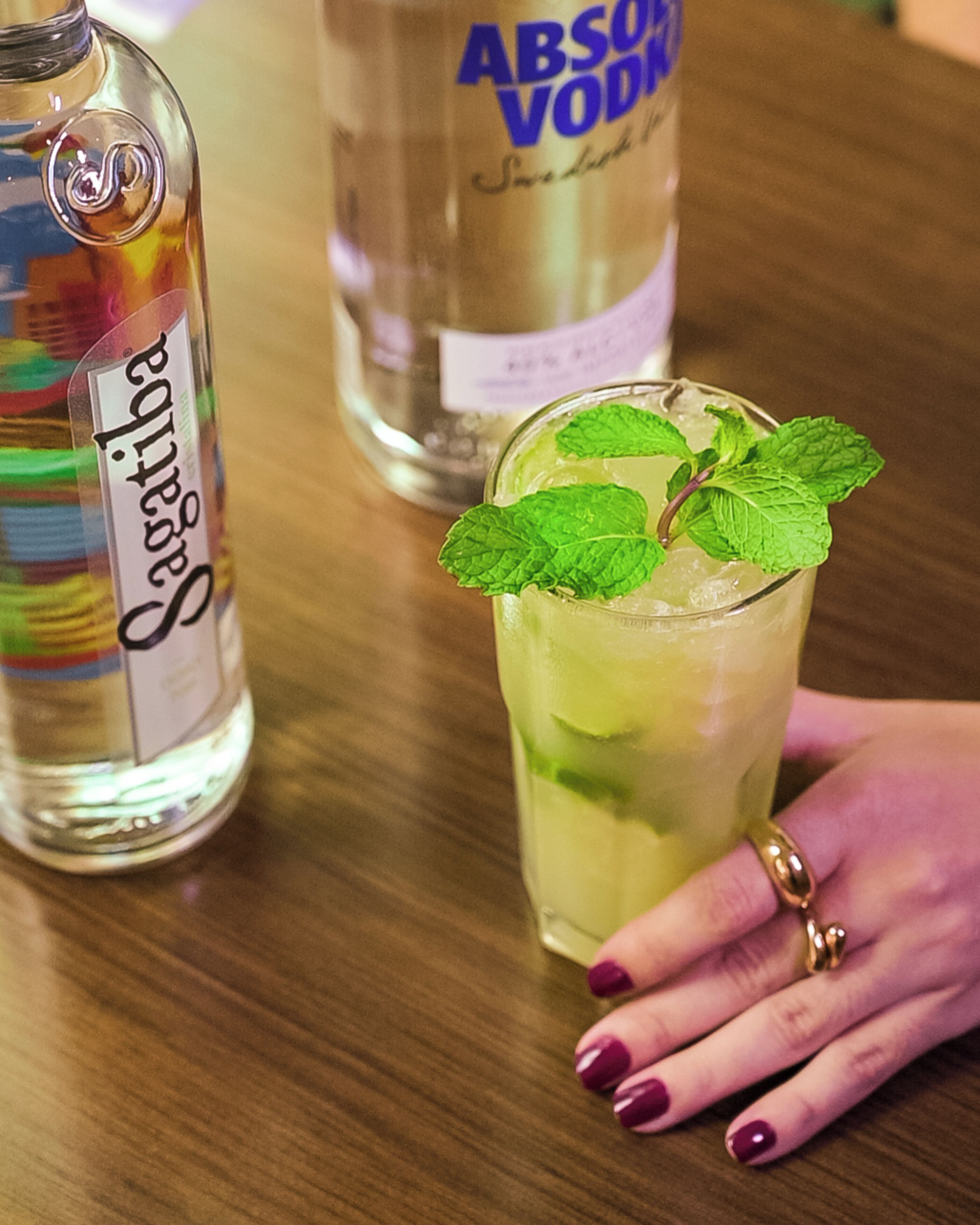Un vaso de una bebida verdosa con hojas de menta, una botella de Absolut Vodka y una botella de Cointreau sobre una mesa de madera.