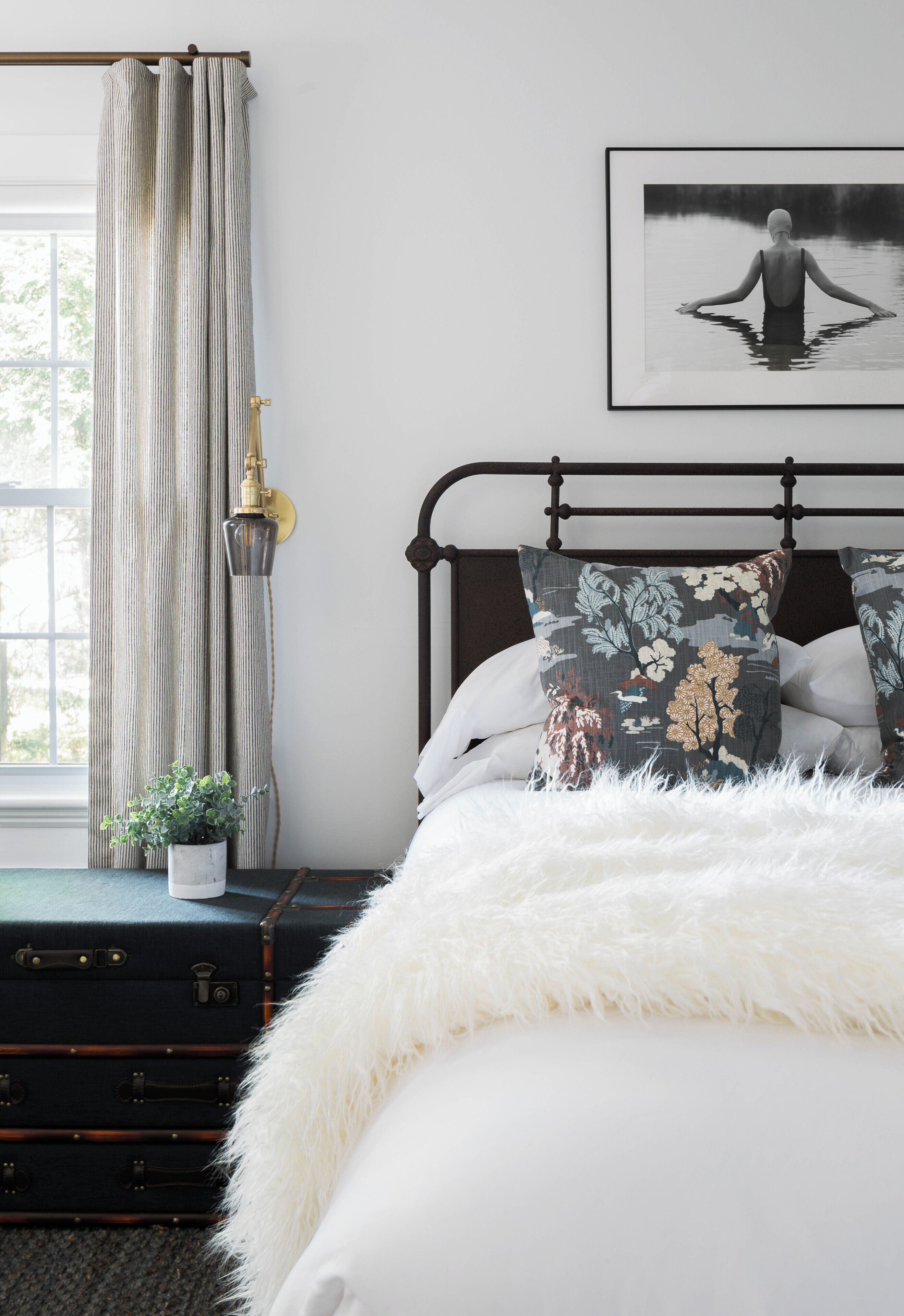 Un dormitorio con cama, una mesita de noche con lámpara, una ventana con cortinas, y un cuadro en la pared.