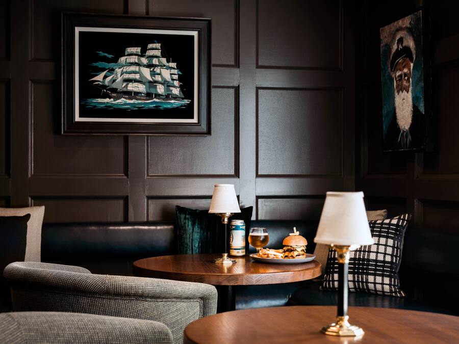 A dark-themed room with a framed maritime painting, a round wooden table with a burger, a beer, and a can of beer, and a sofa with patterned cushions.