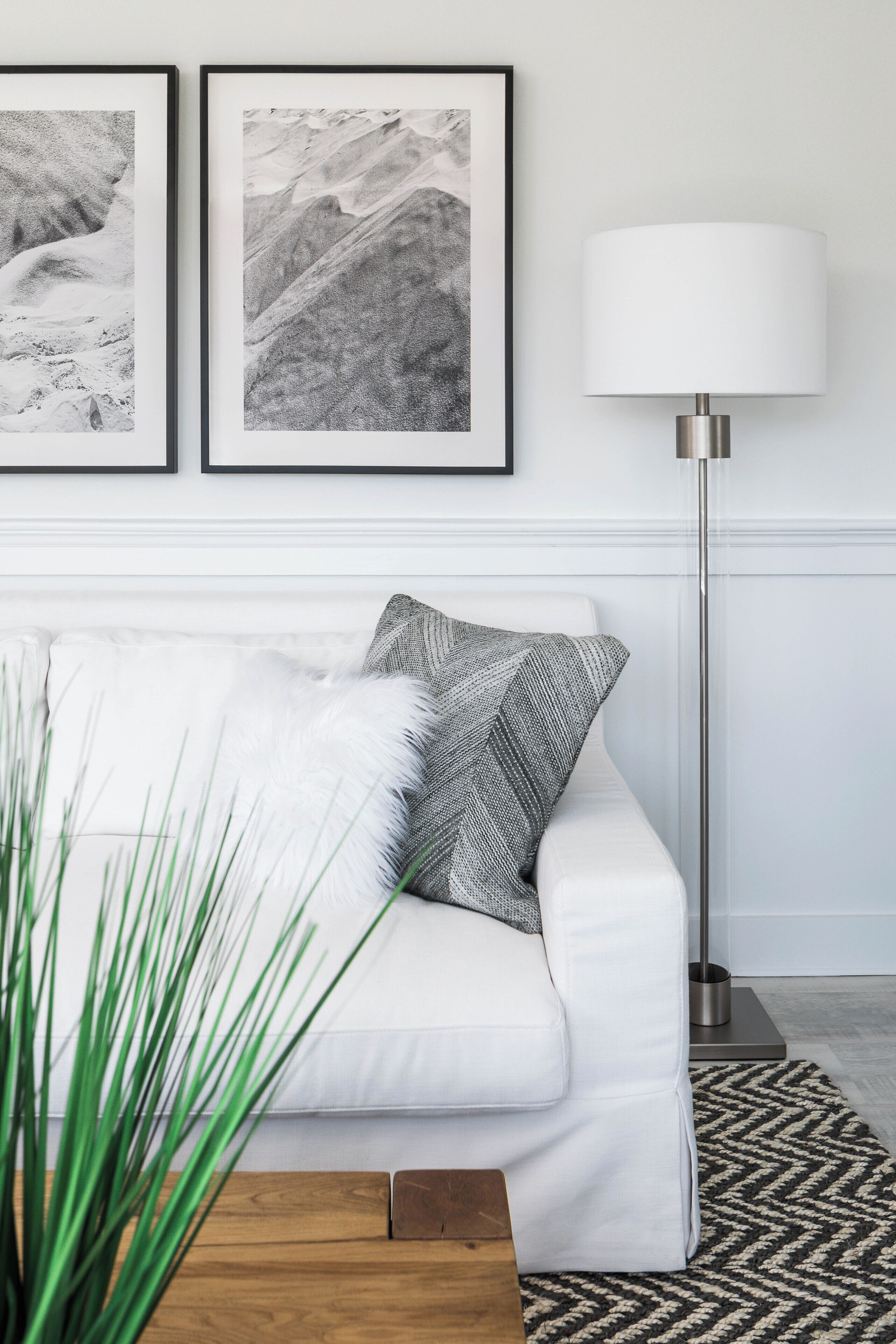 A white sofa with a fluffy pillow and a patterned pillow, a floor lamp, and two framed black and white photographs on the wall.