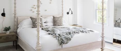 Una cama con dosel de acabado blanco, una mesita de noche con lámpara, y una ventana con vistas al verdor.