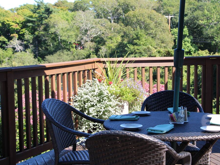 A wooden deck with wicker chairs and a table set for dining, overlooking a lush garden and trees.