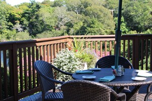 A wooden deck with wicker chairs and a table set for dining, overlooking a lush garden and trees.