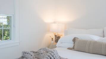 A bedroom with a large bed, a bedside lamp, and a window with a view of greenery.
