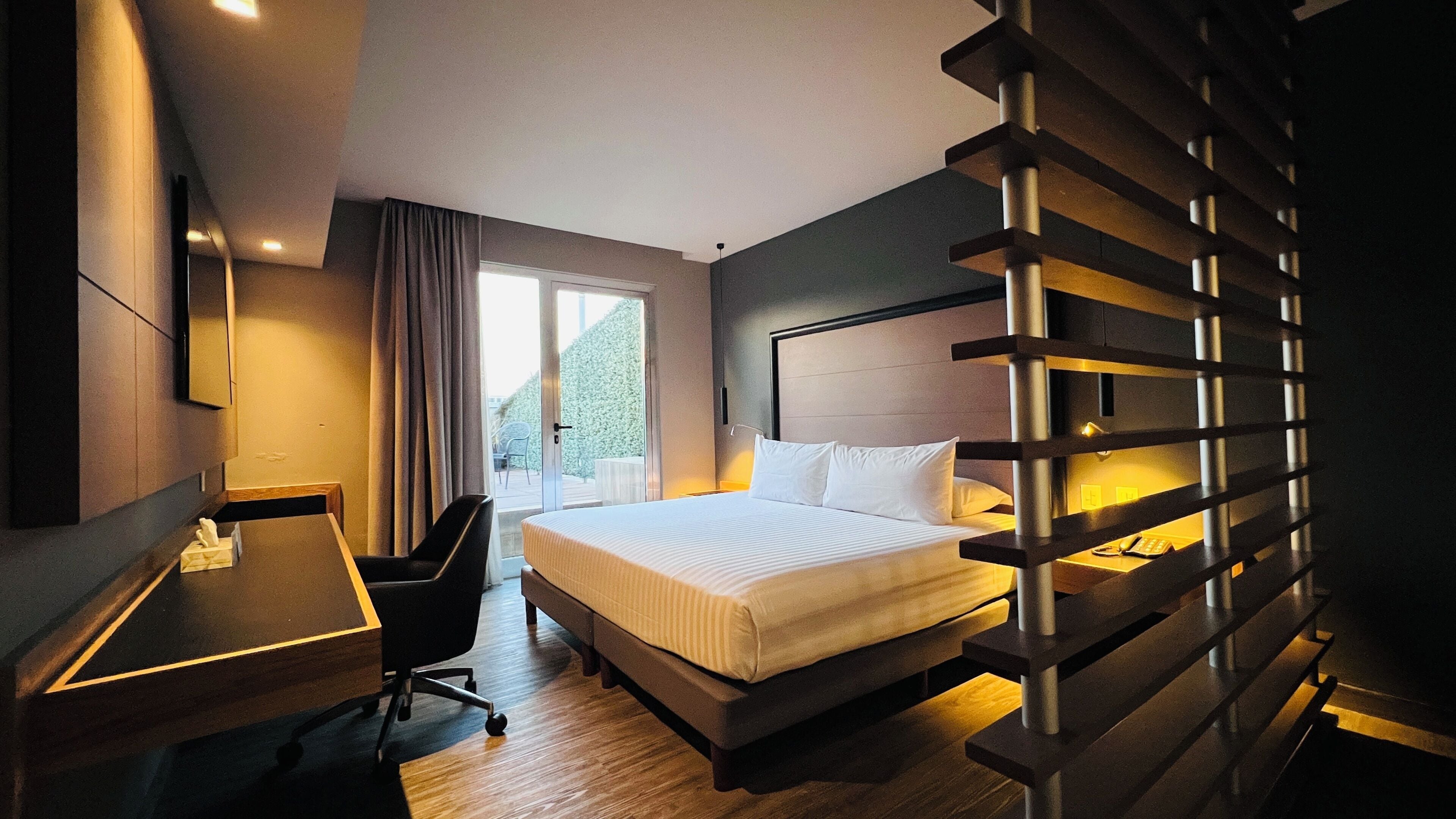 A hotel room with a bed, desk, chair, and a view of the outdoors.