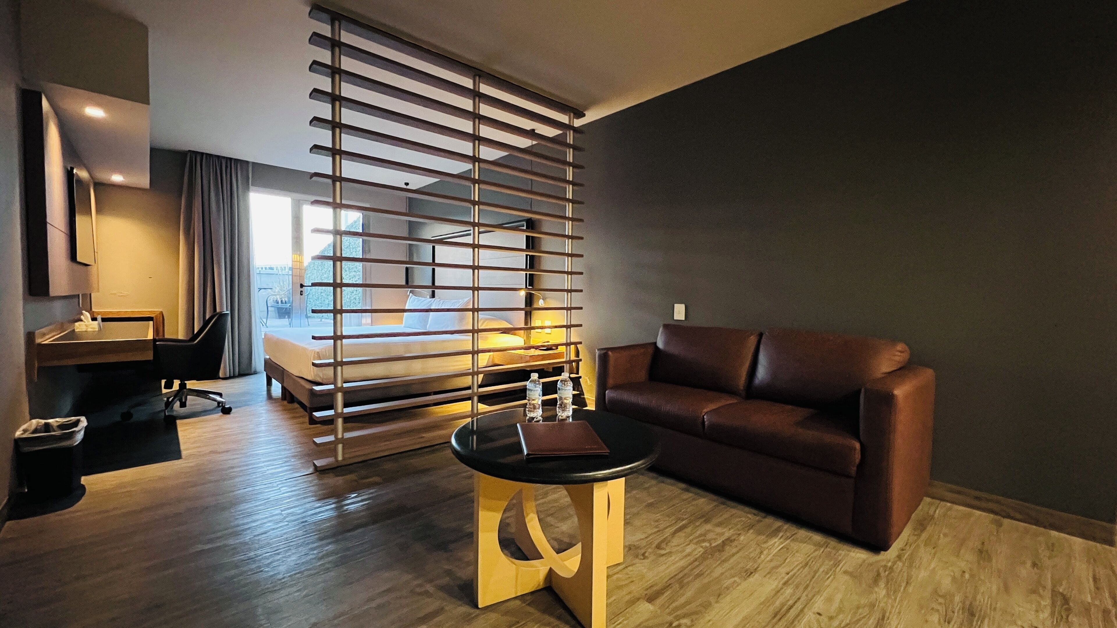 Una habitación de hotel moderna con una cama, un sofá de cuero marrón, una pequeña mesa redonda, un escritorio con una silla y un ventanal con persianas.
