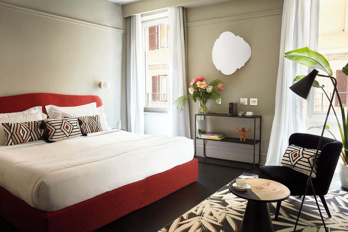 Habitación de hotel con una cama grande, una mesita de noche con lámpara, una silla y vistas al exterior.