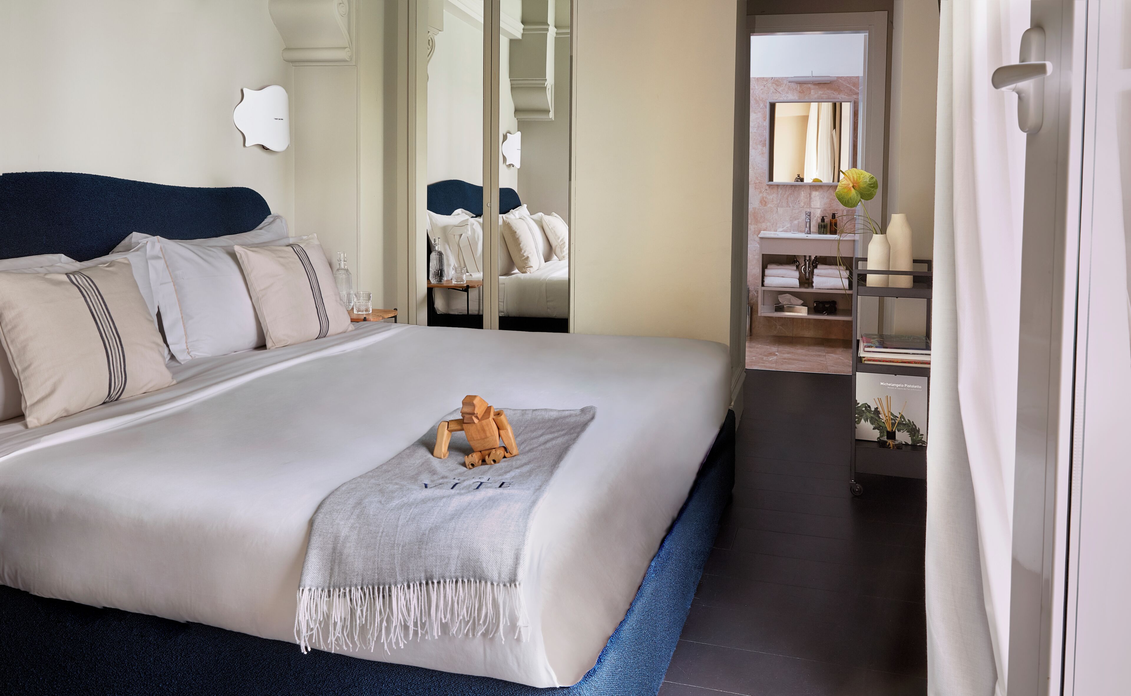 A neatly made bed with a decorative pillow, a bedside table with a glass container, and a mirror reflecting the room.