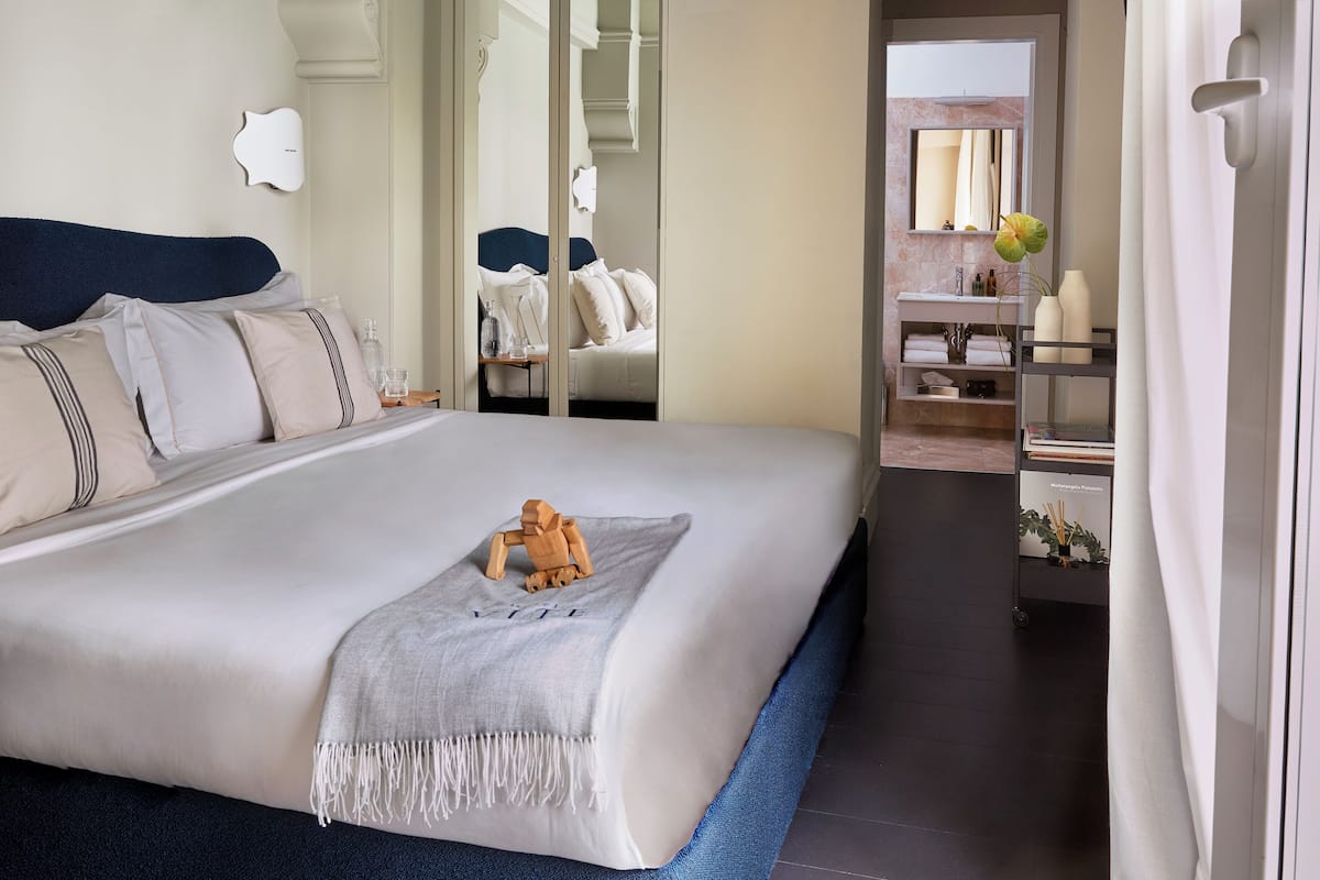 Una habitación de hotel moderna con una cama grande, una mesita de noche de madera, un espejo y un baño con tocador y estantes.