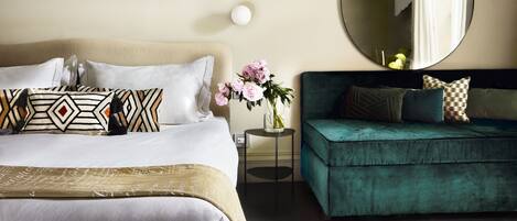 A hotel room with a bed, a sofa, a round mirror, and a vase with flowers.