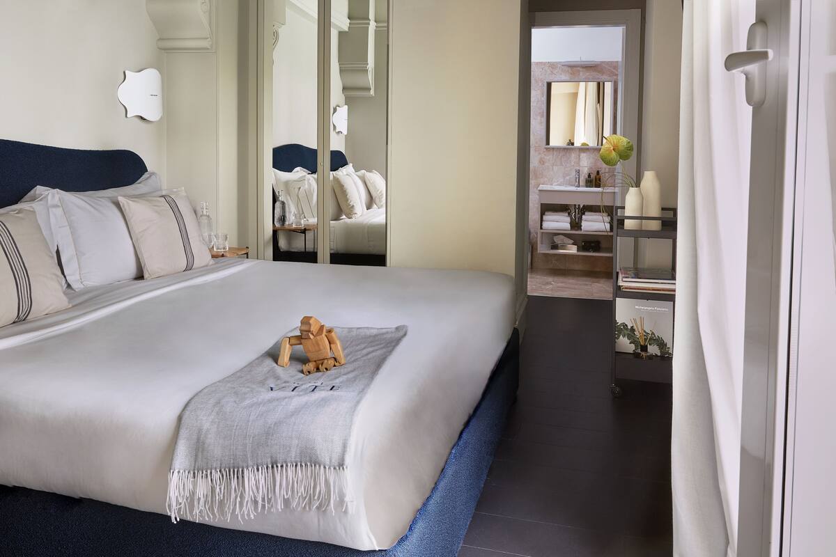 Una habitación de hotel moderna con una cama grande, una mesita de noche de madera, un espejo y un baño con tocador y estantes.