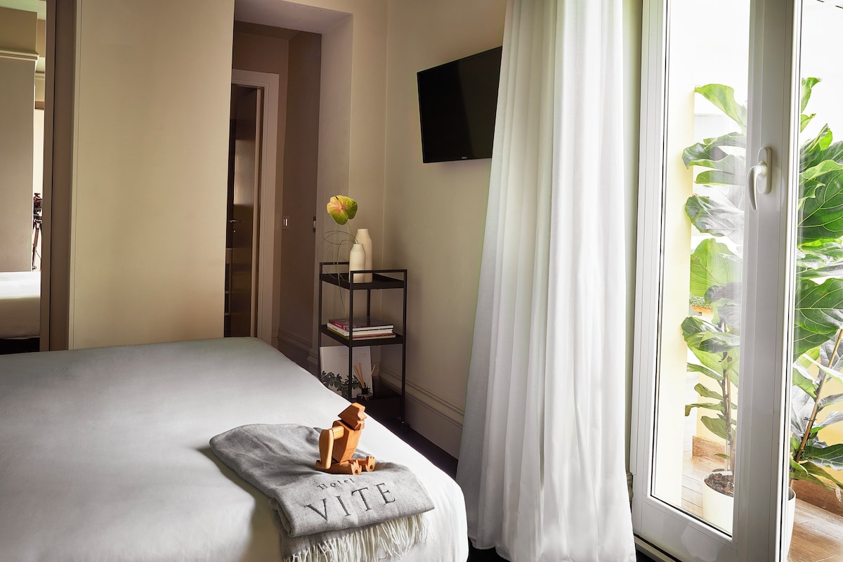 Hotellihuone, jossa on sänky, seinälle asennettu televisio, pieni hylly kirjoineen ja maljakko sekä parveke, jossa on kasveja.