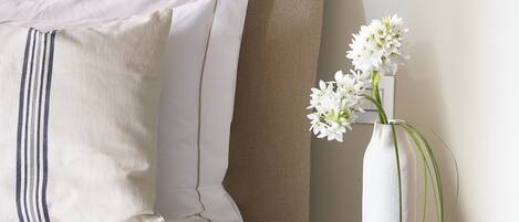 Una cama bien hecha con sábanas blancas y almohadas a rayas, un jarrón blanco con flores blancas sobre una mesita negra, y una escultura de madera en la mesita.